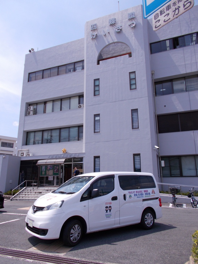 白のミニバンで車体に「特殊詐欺被害防止啓発活動」の内容がラッピングがされた「ふれあい号」が4階建ての四條畷警察署前に停まっている写真