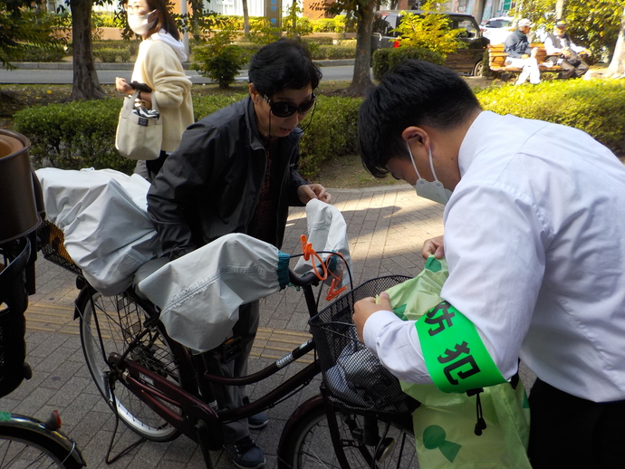 ワイシャツに緑の腕章を付けた男性が年配の女性の自転車のカゴにひったくり防止カバーを付けようとしている写真