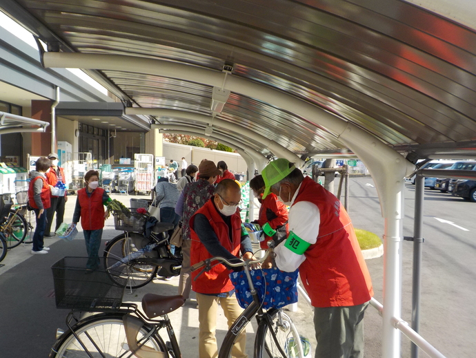万代堺土師店で、赤いベストを着た方々が買い物客の自転車カゴにひったくり防止カバーを取り付けている写真