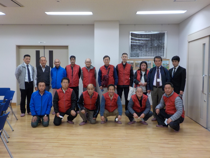 青いジャンバーを着た男性2名と10名の赤いベストを着た八田荘校区自治連合会」の皆さん方と意見交換会で記念撮影をしている写真