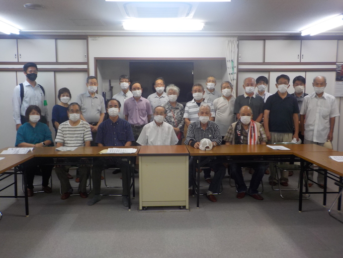 大きなモニターが設置された室内で、八田荘西校区自治連合会の方々と関係者の方々が前列は椅子に座り、後列は立っている集合写真
