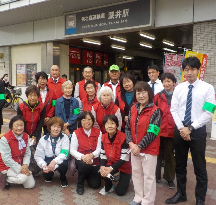 泉北高速鉄道深井駅の前で、左腕に緑の腕章や赤いベストを着た方々と関係者の方々が立っている集合写真