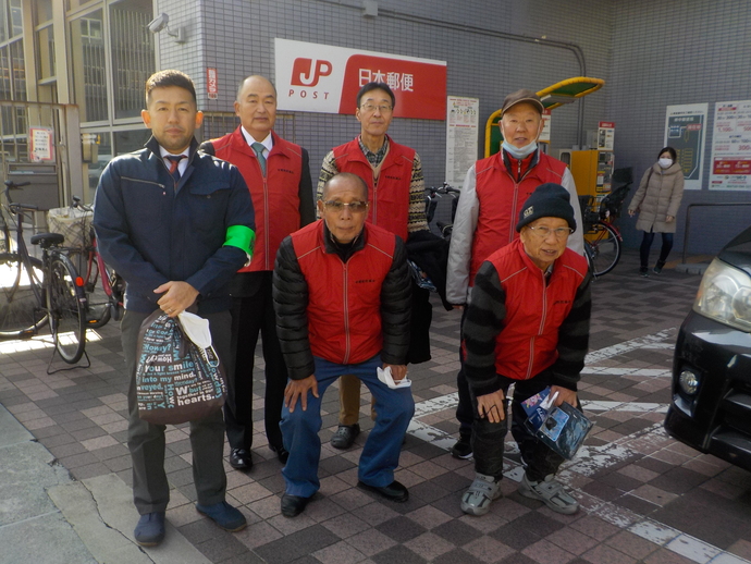 郵便局前でお揃いの赤いベストを着た男性5名と緑のワッペンをつけた男性が記念撮影をしている写真