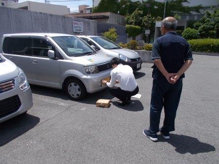 泉南警察署駐車場内で男性がナンバープレート盗難防止ネジの取付をし、車の持ち主の男性が見守っている写真
