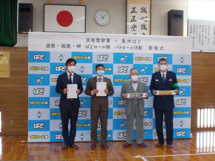 左側の2名の防犯パトロール隊の男性が委託状、右側の2名の男性が腕章を持って記念撮影をしている写真