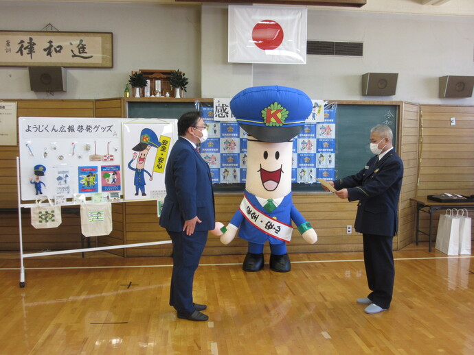 日本国旗が掲げられた広い部屋で、ようじくんが見守る中、河内長野事業場防犯協会の関係者が河内長野警察署長から感謝状を贈呈される様子の写真