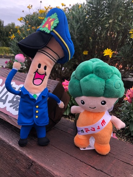屋外の花壇の上に、ようじくんとモックル君のマスコット人形が並んで立てられている写真
