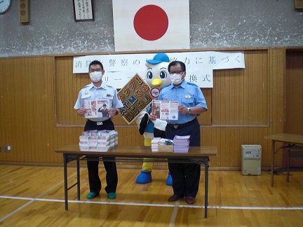 日本国旗が掲げられた部屋で、つるまるくんと2名の警察官が資料が置かれた長テーブルの前に立って並んでいる写真