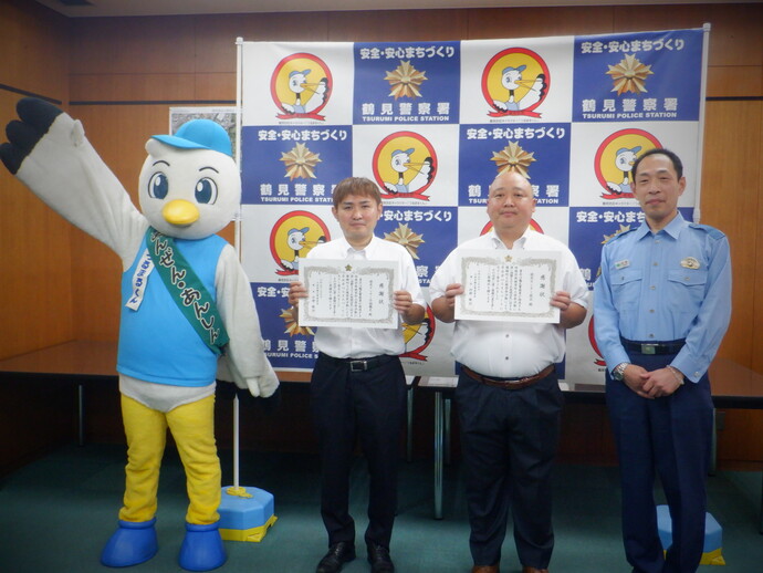 鶴見警察署のバックボードの前で、鶴見防犯協会のマスコットキャラクター「つるまるくん」と警察官のかたの間に2名の男性が立ち、感謝状を持って記念撮影をしている様子の写真