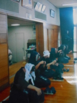 室内の端に一列に並んで座った剣道着姿の人たちが面をつけようとしている様子を写した写真