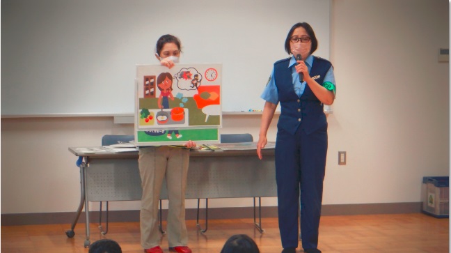 赤いスリッパを履いた女性が5時を過ぎても帰ってこない子どもを心配している母親のイラストを持ち、女性警察官がマイクを左手に持って防犯教室を行っている写真