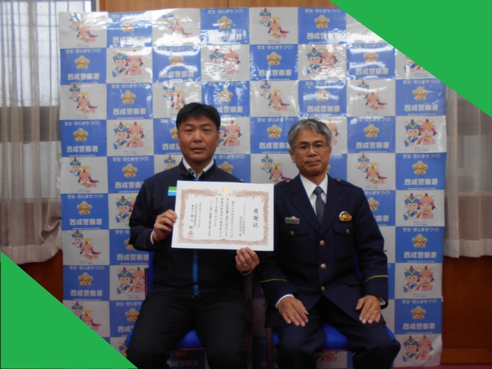 左側に感謝状を持ったファミリーマート岸里駅西店の従業員の男性、右側に警察署員が座っている記念写真