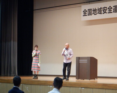 舞台上にマイクを持って立っている一人の女性とタレントのタージンさんの写真