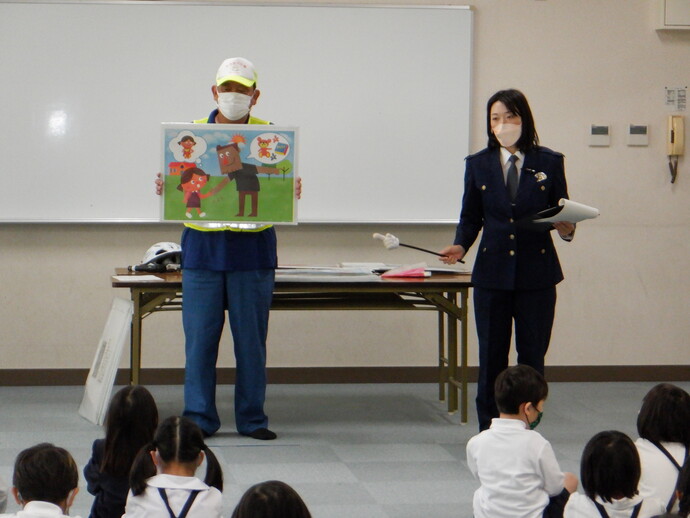 蛍光色の黄色い反射ベストを着た男性が誘拐に巻き込まれそうになっている女の子のイラストを持ち、女性警察官が指差し棒を使って防犯教室を行っている写真
