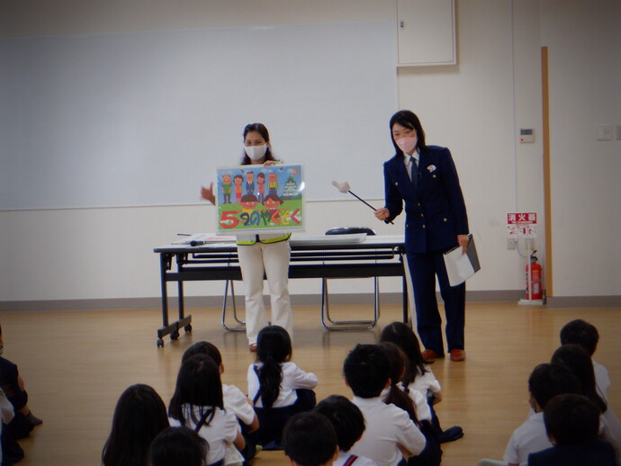 蛍光色の黄色い反射ベストを着た女性が「5つのやくそく」と書かれたイラストを持ち、女性警察官の方が指差し棒を使って防犯教室を行っている写真