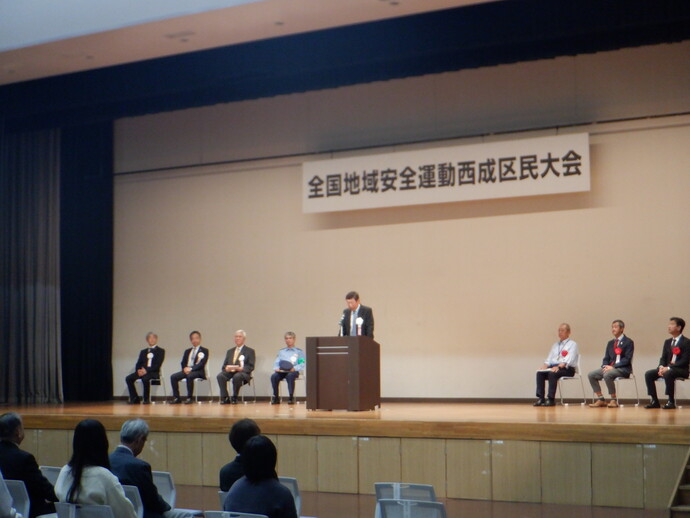 舞台上の演壇に立って話をしている男性、演壇の両脇に並べられた椅子に座っている区民大会参加者の写真