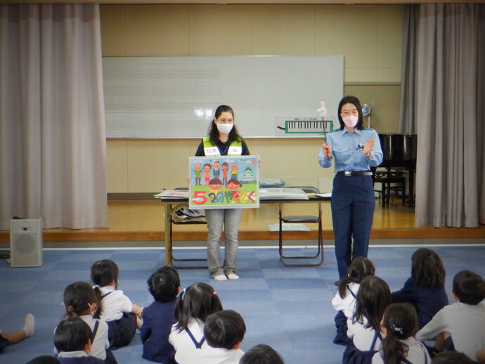 蛍光色の黄色い反射ベストを着た女性が「5つのやくそく」と書かれたイラストを持ち、女性警察官が右手に指差し棒を持ちながらジェスチャーを交えて防犯教室を行っている写真
