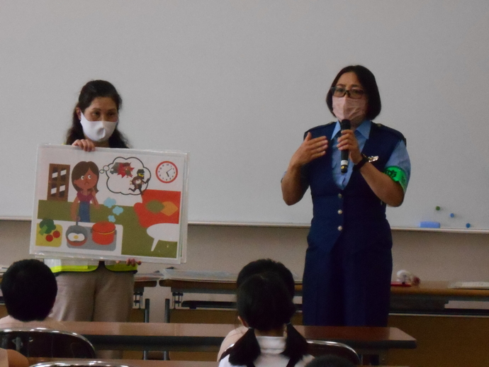 蛍光色の黄色い反射ベストを着た女性が5時を過ぎても帰ってこない子どもを心配している母親のイラストを持ち、女性警察官がマイクを左手に持ちながらジェスチャーを交えて防犯教室を行っている写真