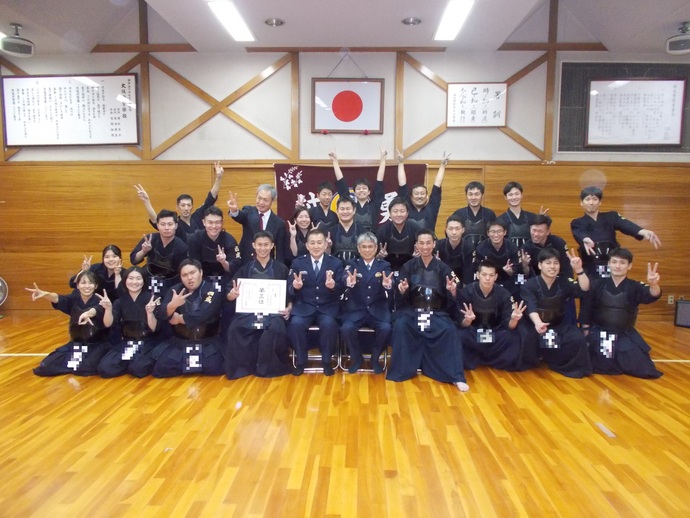 中央の椅子に座った制服姿の男性2名の周りに剣道着姿の方々が手でピースサインをしている集合写真
