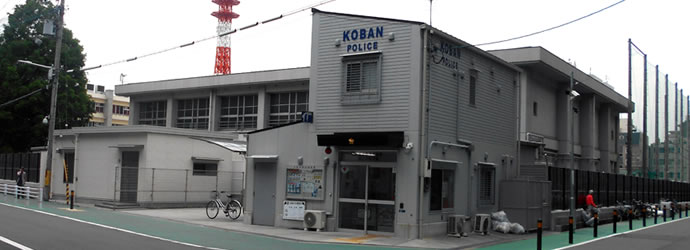 後方に見える学校に隣接された壁が灰色で片流れ屋根の2階建てになっている萩之茶屋交番の外観写真
