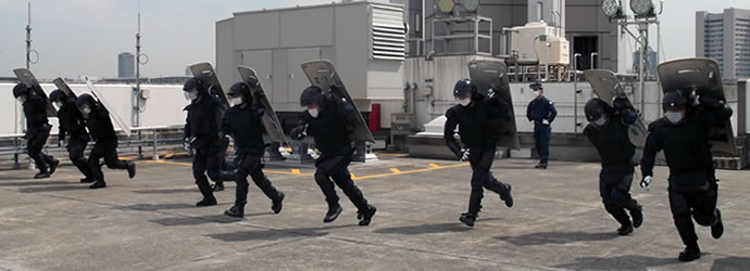 9人の警察隊が防護服とヘルメットをかぶり、防護盾を左手で担いで訓練している写真