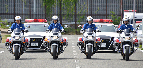 交通機動隊のバイク4台が横並びで走り、その後ろにパトカー2台が追走している写真