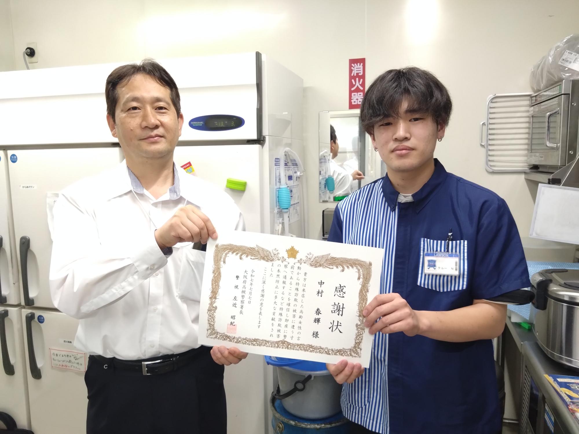高槻野田郵便局の女性局員が感謝状を手に持ち、郵便局員や緑色の腕章をつけた男性と横1列に並んでいる写真