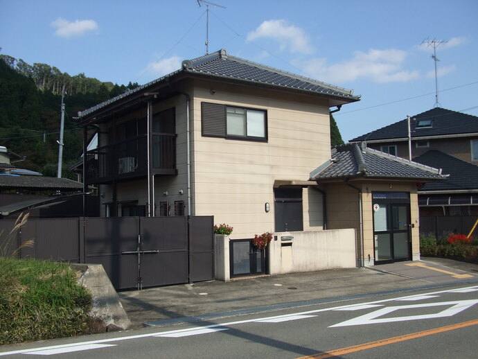 外壁がクリーム色の瓦屋根の2階建てでベランダがあり、建物横には奥が見えづらい大人の背丈をほどの黒色の門扉が設置されたスペースと駐在所の玄関とは別に住居用の玄関がある今西駐在所の外観写真