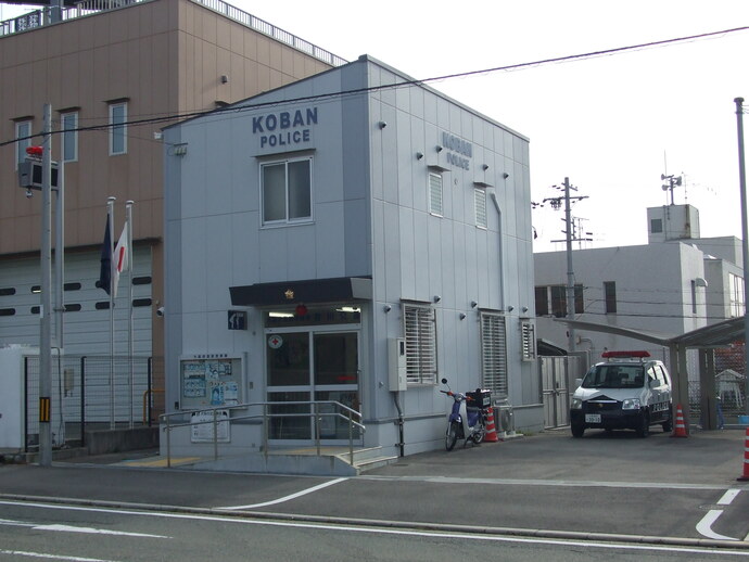 外壁がグレー色の2階建てで入口には手すり付きのスロープがあり、建物右側に駐車スペースとパトカーが停められている吉川交番の外観写真