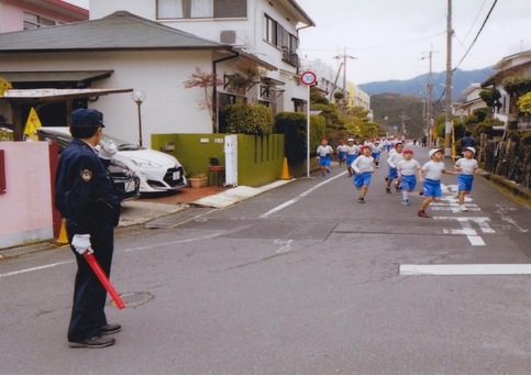 体操服姿の園児達がマラソン大会で道路を走っている様子を駐在所勤務員が交通整理をして見守っている様子を写した写真