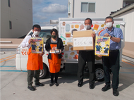 防犯啓発品が入った段ボールやポスターを持っている、オレンジ色のエプロンをつけた店員と警察署員4名が「移動スーパー・とくし丸」の車両の前で記念撮影をしている写真