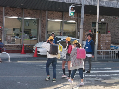 黄色の帽子を被った小学生3人が横断歩道をわたるところを見守り隊の男性2名が見守っている写真