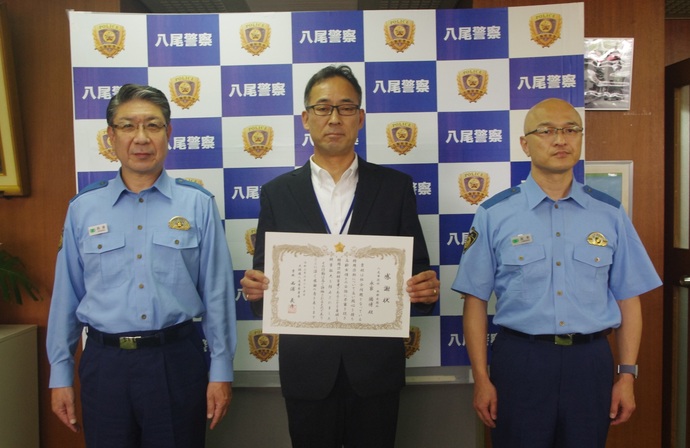 八尾警察のバックボードを背景に感謝状をもったスーツ姿の男性と両隣に警官服を着た男性2人が記念撮影をしている写真