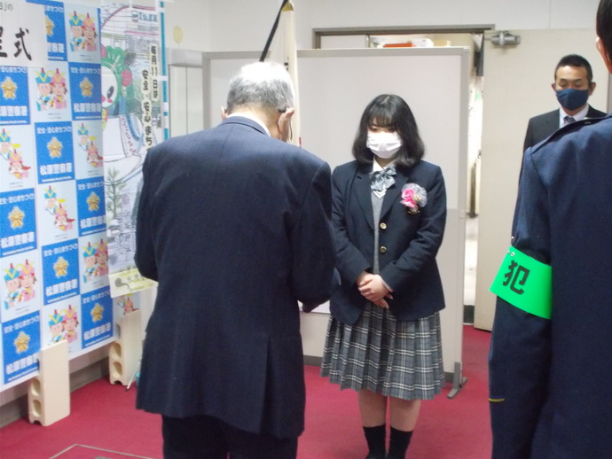 「毎月11日は安全・安心まちづくりの日」に掲げる啓発のぼり旗が置かれ、制服を着た女子学生が立ち、スーツ着た男性が感謝状を読んでいる贈呈式の写真