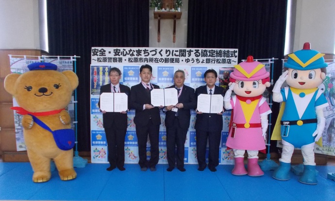 安全・安心まちづくりに関する協定締結式と書かれたバックボードの前に4人の関係者が協定書を開いて持ち、左側に日本郵便のキャラクターぽすくま、大阪府警のキャラクターフーくん・ケイちゃんが敬礼した記念撮影の写真
