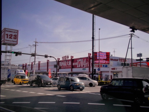 春の地域安全運動に関する広報映像が映し出された123+N松原店の電光掲示板を車の通る大通りから写した外観写真