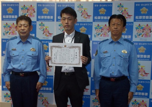 職員の男性が賞状を持ち、両隣に警官が並んで記念撮影をしている写真
