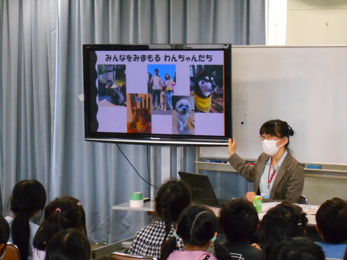 犬の写真が複数映し出されたモニターに手を添えて、目の前に座る子ども達に話をしている女性の写真