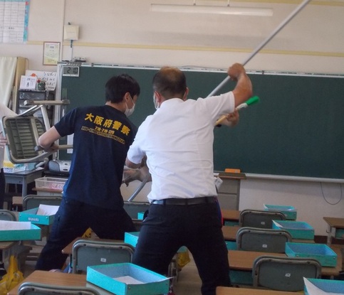 さすまたを持った男性2名が教室内の不審者を押さえつけている訓練の写真