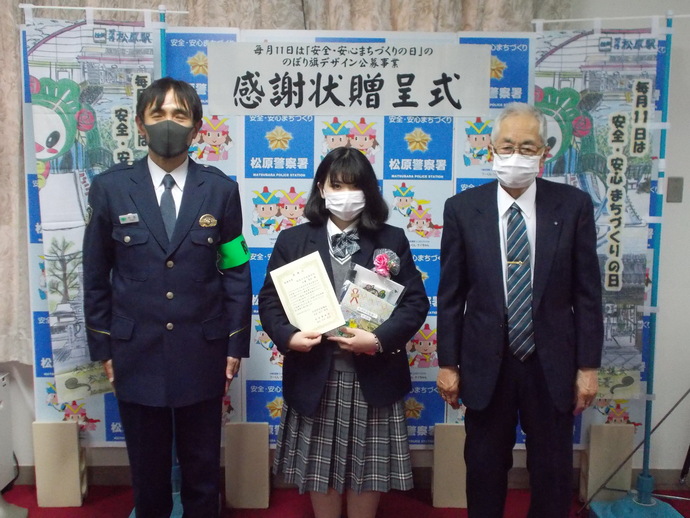 感謝状贈呈式と書かれ、「毎月11日は安全・安心まちづくりの日」に掲げる啓発のぼり旗が両側に置かれ中央に阪南大学高等学校3年生の生徒、左に警察署員、右にスーツの男性が並び3人で撮影した記念写真