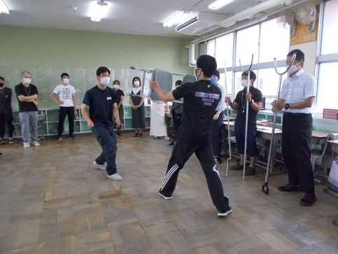 窓際にさすまたを準備して立っている3名の教師と、中央で椅子を使い犯人と応戦している先生の訓練を教室内で行っている写真