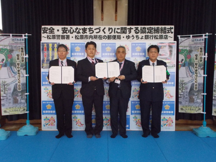 安全・安心まちづくりに関する協定締結式と書かれたバックボードの両側に毎月11日の安全・安心まちづくりの日の、のぼり旗が置かれ、前に4人の関係者が協定書を開いて持って記念撮影している写真
