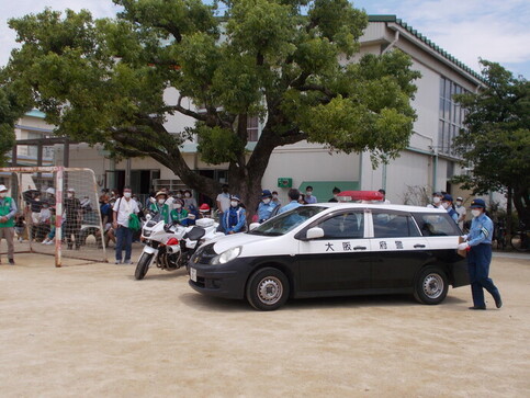 子供たちが体育館の大きな木の前に停められたパトカーと白バイを見学している写真
