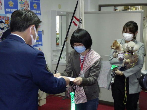 警察官より女性が委嘱状を受け取り、その後ろの女性が小型犬を2匹、抱っこしている写真