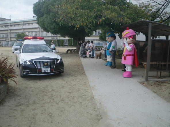 小学校の校庭をパトロールカーが出発し、その横でフーくん・ケイちゃんが手を振って見送っている写真