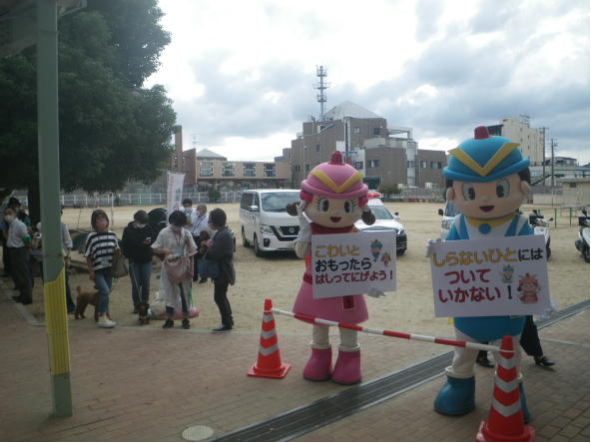 学校の敷地内でケイちゃんが、こわいとおもったらはしってにげよう!と書いたパネルを持ち、フーくんが、しらないひとにはついていかない!と書いたパネルを持ち、左側に松原わんわんパトロール隊が集まっている写真