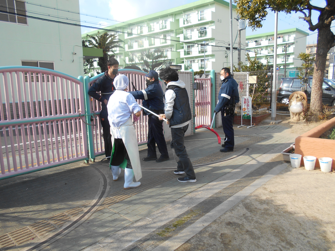 保育園の門で白衣を着てエプロンと長靴をはいた職員と男性の職員で、不審者役をさすまたを使い捕らえる指導を受けている写真