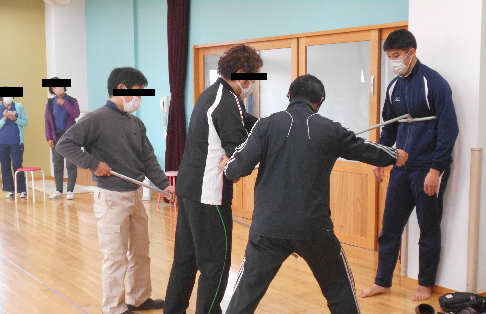 さすまたを持った男性2名のうち1人が、ジャージ姿の男性をさすまたを使い壁に押さえつけている不審者対応訓練の写真