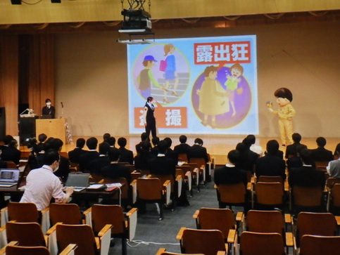 ステージに設置されたスクリーンに階段を上る女性のスカートの中を盗聴している様子と男性がコートを広げ女性が慌てている露出狂のイラストが映し出され、阪南大学高等学校の学生たちが椅子に座って聞いている防犯教室の様子を後ろから撮影した写真