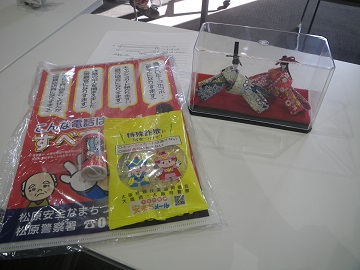 高齢者参加のハンドメイド教室で作成されたお雛様と配布された、松原警察署オリジナル特殊詐欺被害防止啓発チラシと安全安心まちづくりしんちゃんラムネの写真